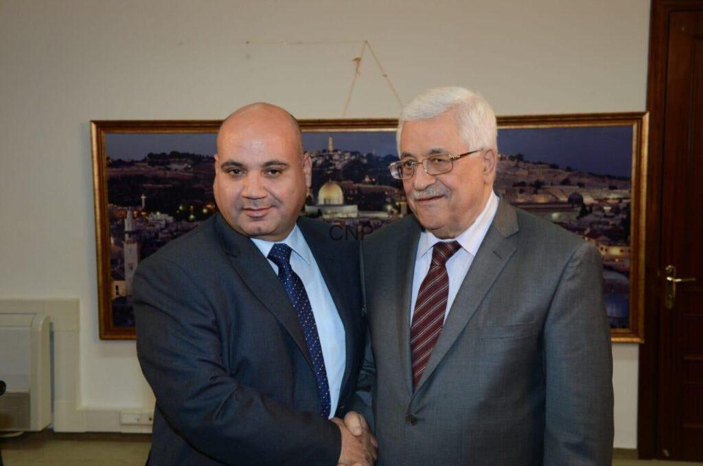 Two men in suits shake hands in front of a framed cityscape photo in an office setting.