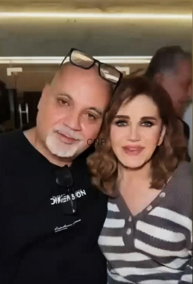 Two smiling people posing for a selfie indoors, man with glasses on his head and a woman beside him in a striped top.