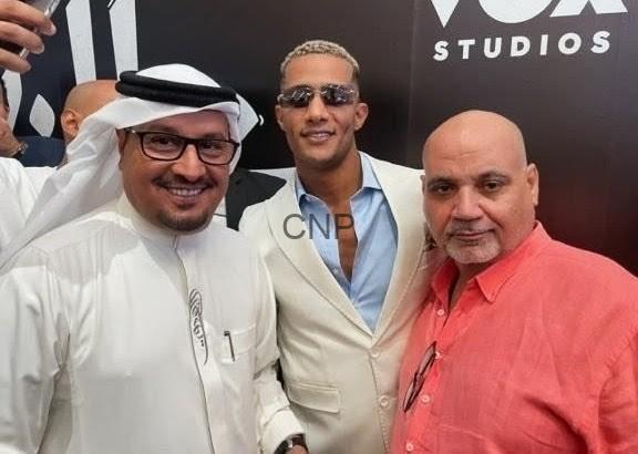 Three men pose for a photo at a Vox Studios event: left in white traditional attire and glasses, middle in a beige blazer with sunglasses, right in a coral shirt.