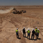 Construction crew in hard hats and neon vests observe desert earthworks as heavy machinery dig and move soil in a dusty excavation site.