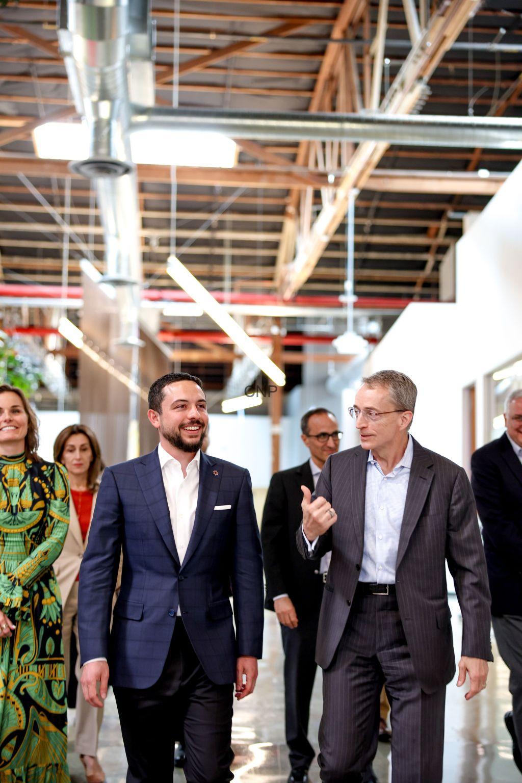 Group of professionals walking and chatting in a bright, open office with exposed ceilings and ductwork. A man in a blue suit smiles as he walks beside a gray-suited colleague who gestures while speaking.