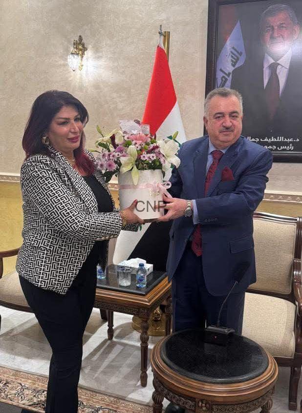 Man in a blue suit hands a white flower pot to a woman in a patterned blazer as they stand in a formal room with a red-and-white flag and a portrait in the background, the bouquet featuring pink flowers.