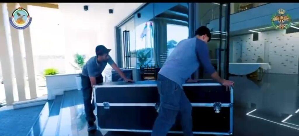 bb2 - Center News Network Two men move a large black flight case across a tiled outdoor area in front of a glass-building entrance, preparing for transport.