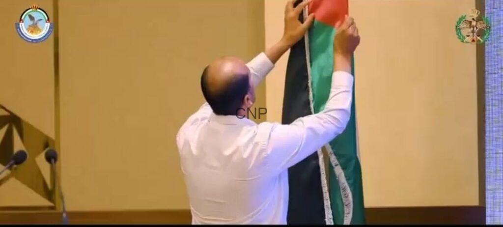 Man in a white shirt raises a tricolor flag (green, black, red) on a stage, with microphones to the left and ceremony decor nearby.