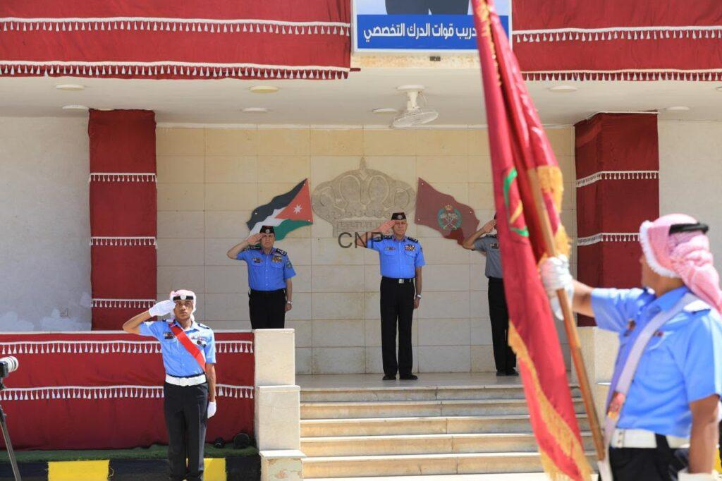 Three air force officers salute on a tiled stage during a ceremony, with a flag bearer in the foreground and a national flag in the background.