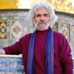 Portrait of an older man with gray hair and beard in a maroon shirt and blue scarf, standing before a colorful mosaic-tiled structure.