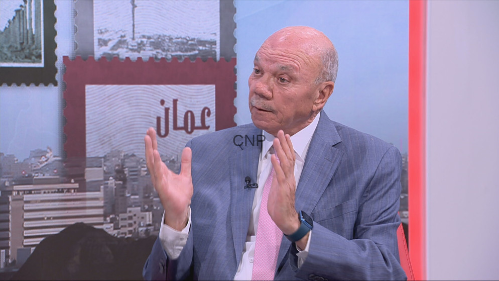 Bald man in a light blue suit and pink tie speaks on a TV set, gesturing with both hands while wearing a lapel mic.