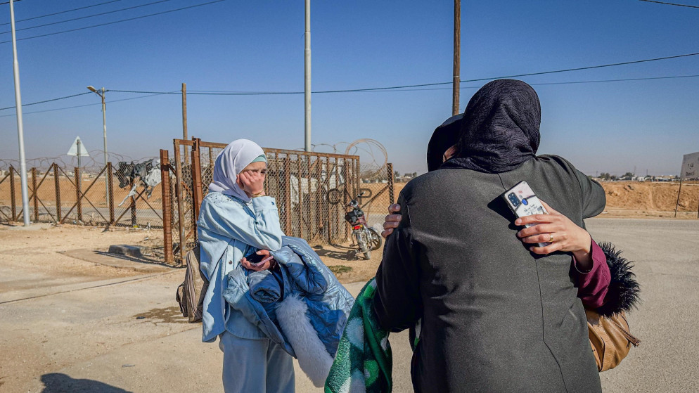 Two women wearing headscarves hug and take a selfie near a fenced border area in a desert setting.