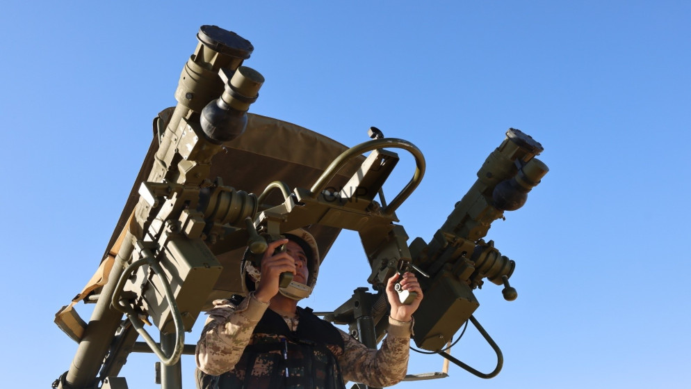 Military service member in desert camouflage operating a large dual-barrel mounted weapons system outdoors under a blue sky