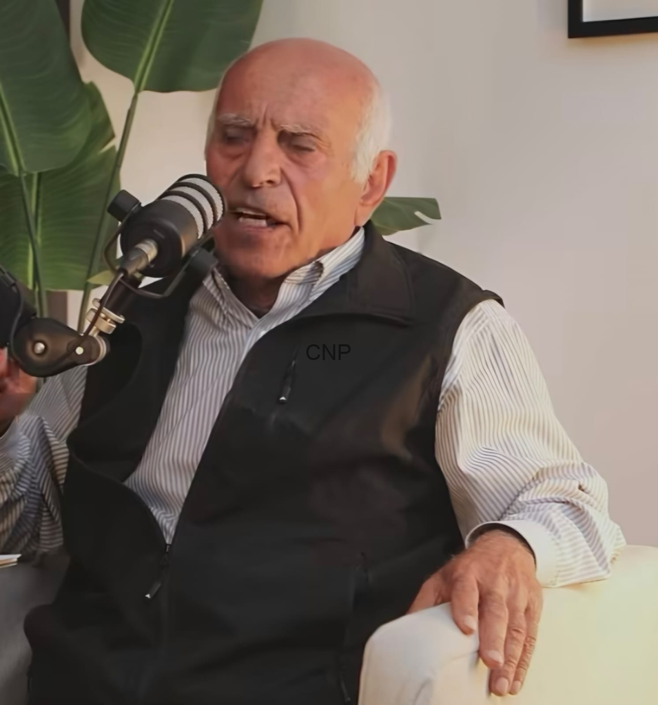 Older man with white hair speaks into a studio microphone during an interview or talk.