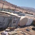 Desert industrial yard with rusted cylindrical tanks and metal containers, ladders, and scattered hoses, with two workers in the foreground and mountains in the distance.