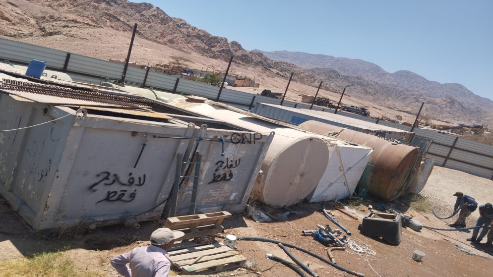 image - Center News Network Desert industrial yard with rusted cylindrical tanks and metal containers, ladders, and scattered hoses, with two workers in the foreground and mountains in the distance.