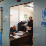 Two men in a small office signing and reviewing documents, with the UNHCR logo on the blue door outside the room.