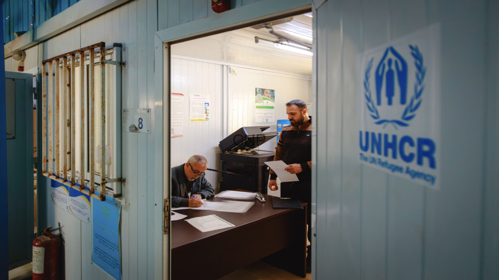 image - Center News Network Two men in a small office signing and reviewing documents, with the UNHCR logo on the blue door outside the room.
