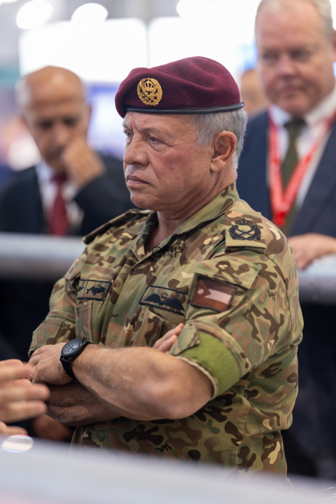 image - Center News Network Older man in camouflage uniform with a maroon beret and a gold insignia, arms crossed, in a crowd scene.