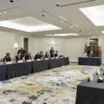 Formal diplomatic meeting in a bright hotel ballroom; delegates sit along two long tables facing each other, Jordan flag at the head table.
