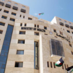 Beige government building with a prominent 'MINISTRY OF HEALTH' sign, two flags (Palestinian and Jordanian) waving in the foreground against a clear blue sky.