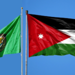 Two flags flying on flagpoles against a clear blue sky: a green flag with a circular emblem on the left and the Jordan flag on the right, with black, white, green stripes and a red triangle.