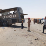 Bus overturned on a desert highway after a crash; bystanders and security personnel assess the scene.