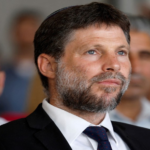 Bearded man in a dark suit and navy tie listening at a formal event, with a blurred audience in the background.