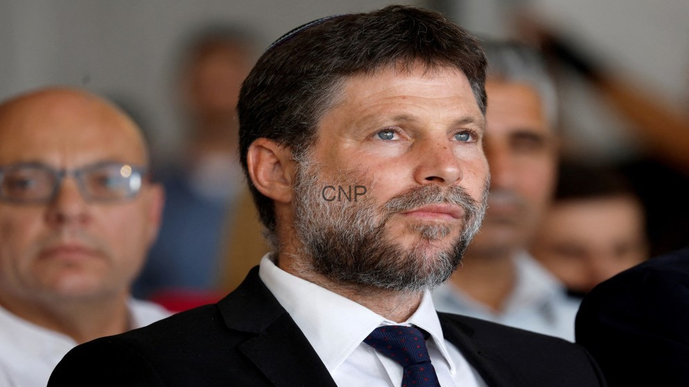 image - Center News Network Bearded man in a dark suit and navy tie listening at a formal event, with a blurred audience in the background.