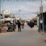 Busy market street in a Middle Eastern town with stalls, vendors, and shoppers along a paved road under string bunting.