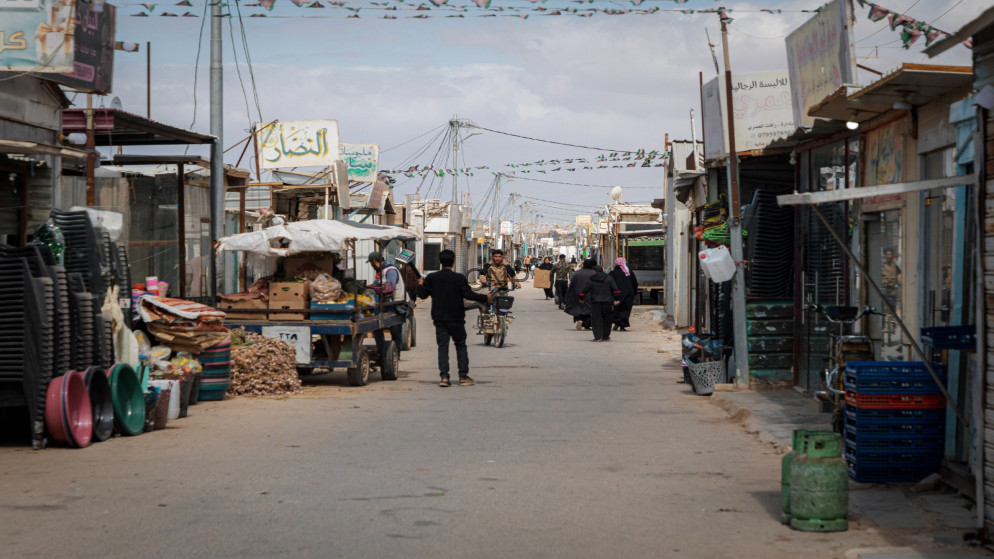 Busy market street in a Middle Eastern town with stalls, vendors, and shoppers along a paved road under string bunting.