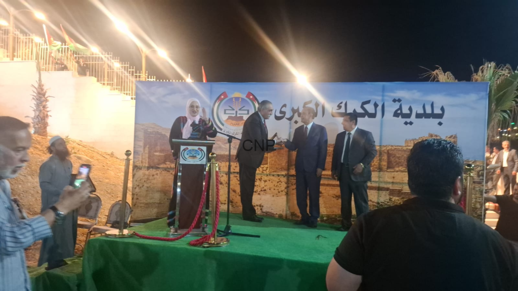 Outdoor night ceremony: two men in suits shake hands on a green stage with a podium and a banner in Arabic behind them, attendees watching.]@