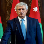Middle-aged man in a navy suit and light blue tie standing at a podium, with Jordanian flags in the background.