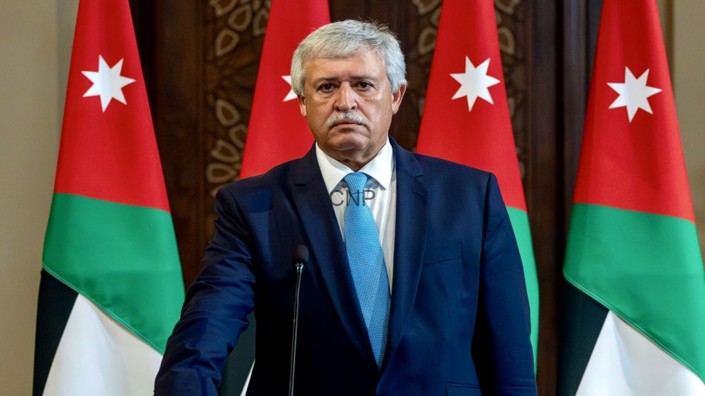 image - Center News Network Middle-aged man in a navy suit and light blue tie standing at a podium, with Jordanian flags in the background.