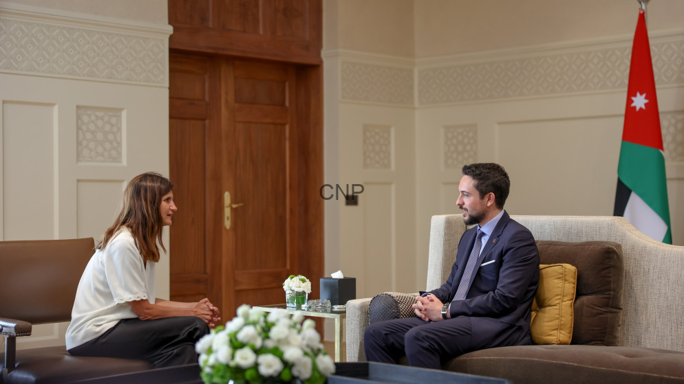 Two people sit facing each other in a formal office: a woman in a white blouse and a man in a navy suit, with a Jordanian flag in the background.