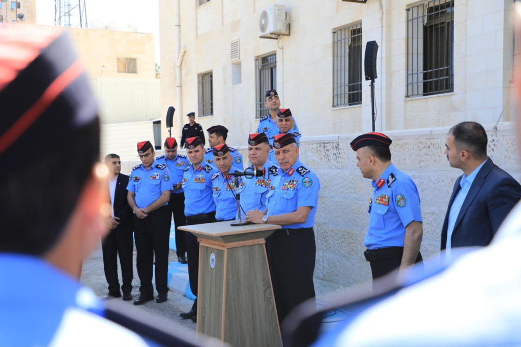 Police officers in blue uniforms stand at a wooden podium giving a speech outdoors, with a row of officials and onlookers behind them in a courtyard building setting