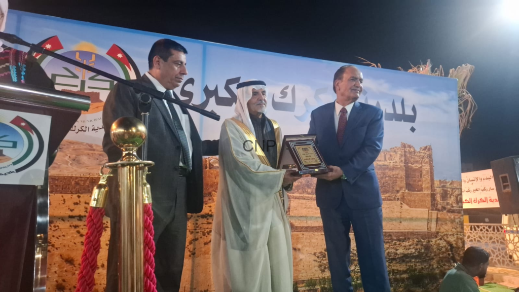 Three men on a stage during an award ceremony; a man in traditional Arab dress hands a plaque to a suited man as another man looks on, with Arabic text on a backdrop behind them.