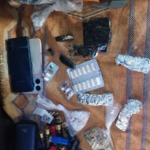 Assorted drug paraphernalia on a patterned cloth: blister packs, pills, foil packets, plastic bags, and a phone nearby.