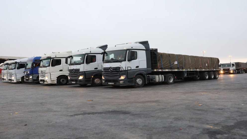 image - Center News Network Row of white semi-trucks parked in a lot, connected to long trailers covered with tarps on a gray day