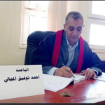 Man in a suit with a red ceremonial sash signs documents at a desk; a sign in Arabic sits in front of him on the table.