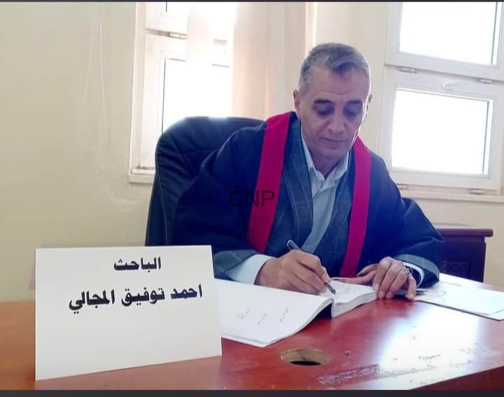 Man in a suit with a red ceremonial sash signs documents at a desk; a sign in Arabic sits in front of him on the table.