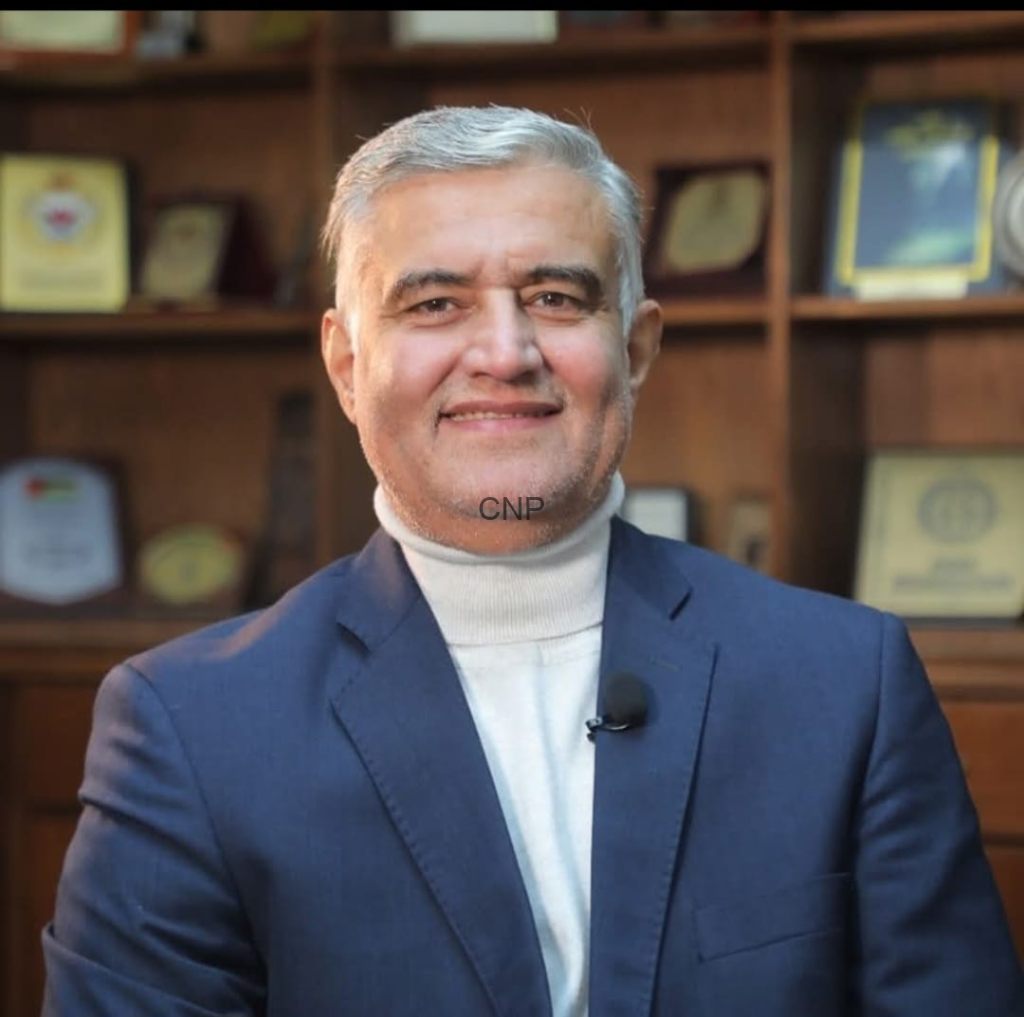 Smiling middle-aged man in a navy suit with a cream turtleneck, mic clipped to his lapel, in an office with wooden shelves and awards in the background.