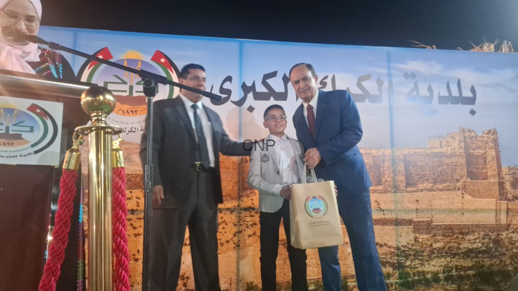 image - Center News Network Boy in a light gray blazer receives a gift bag on a stage, flanked by two men in suits; podium with microphone and red rope in front, backdrop with Arabic text and ruins.