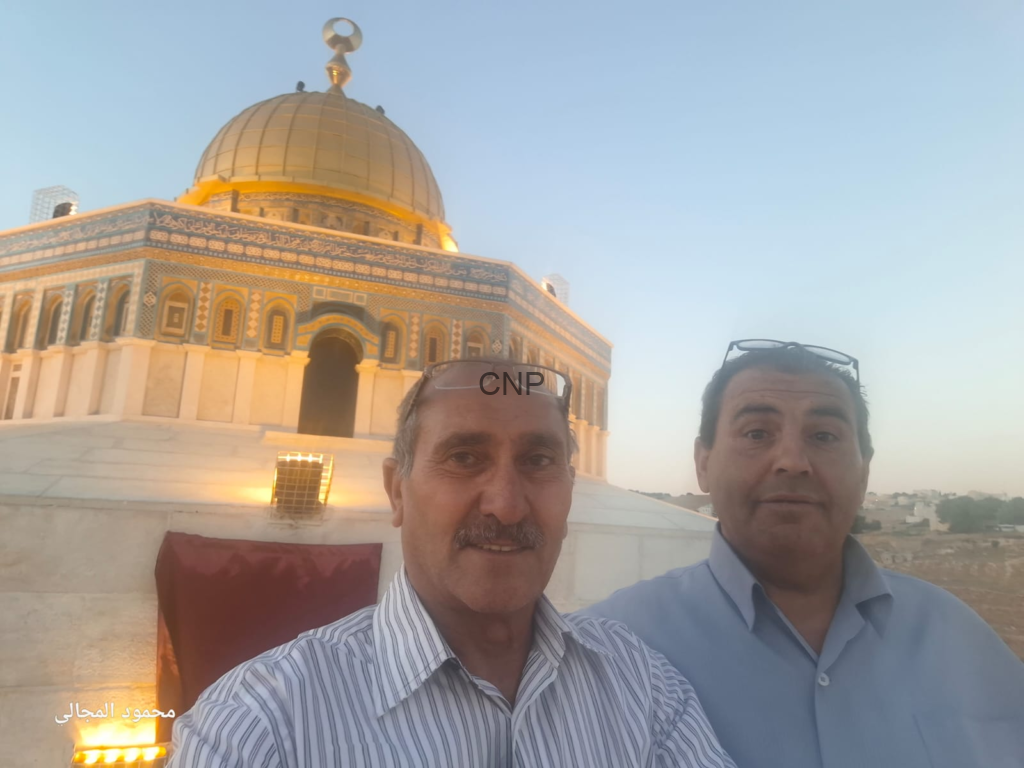 image - Center News Network Two men taking a selfie in front of the Dome of the Rock with its golden dome and decorative façade visible against a clear blue sky.