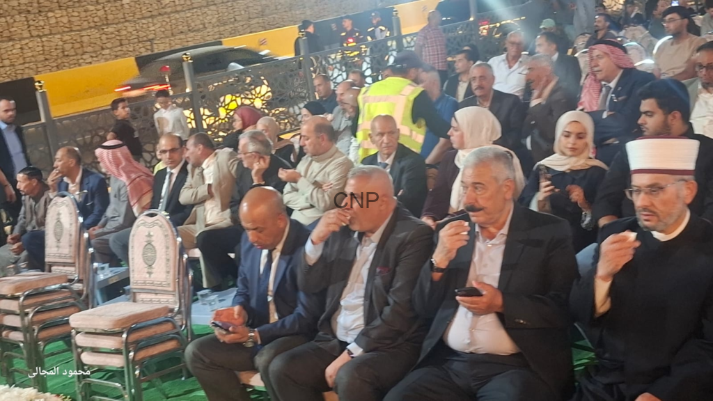 Crowd of men and women seated at an outdoor event; a security guard in a yellow vest stands behind them, some in traditional dress and hijabs, others with phones