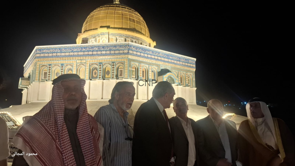 image - Center News Network Nighttime group portrait in front of the illuminated Dome of the Rock, with the gold dome and blue-tiled façade visible behind the men.