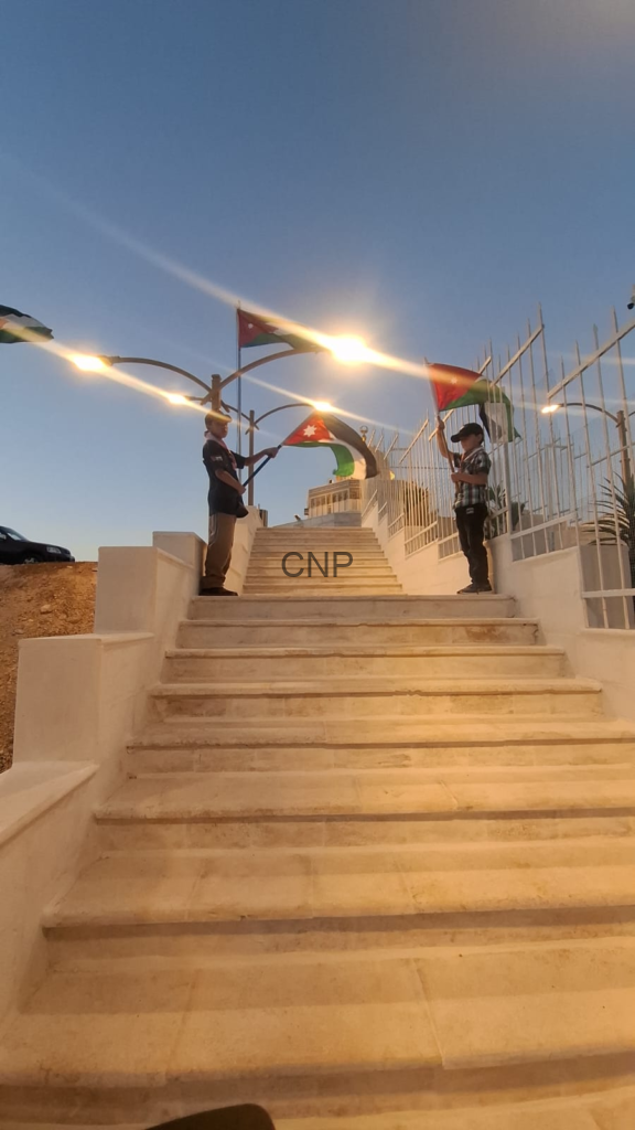 Two people on a white stone staircase hold Jordanian flags against a clear blue sky.