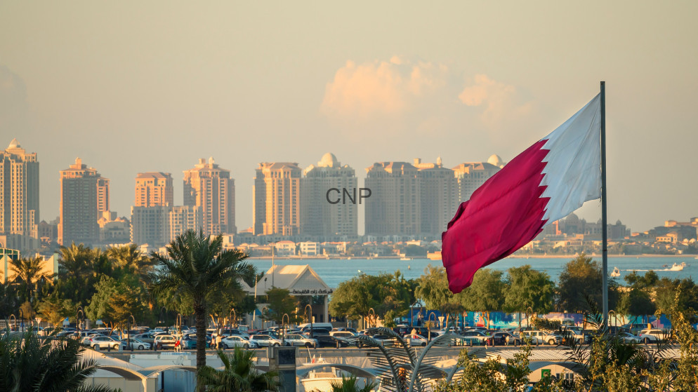 Qatar flag waving beside a waterfront skyline with tall buildings and palm trees in the foreground.