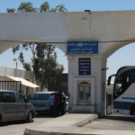 Border checkpoint with arched entrances, a small booth, blue VIP sign, and a white-blue bus on the right with parked vans nearby.