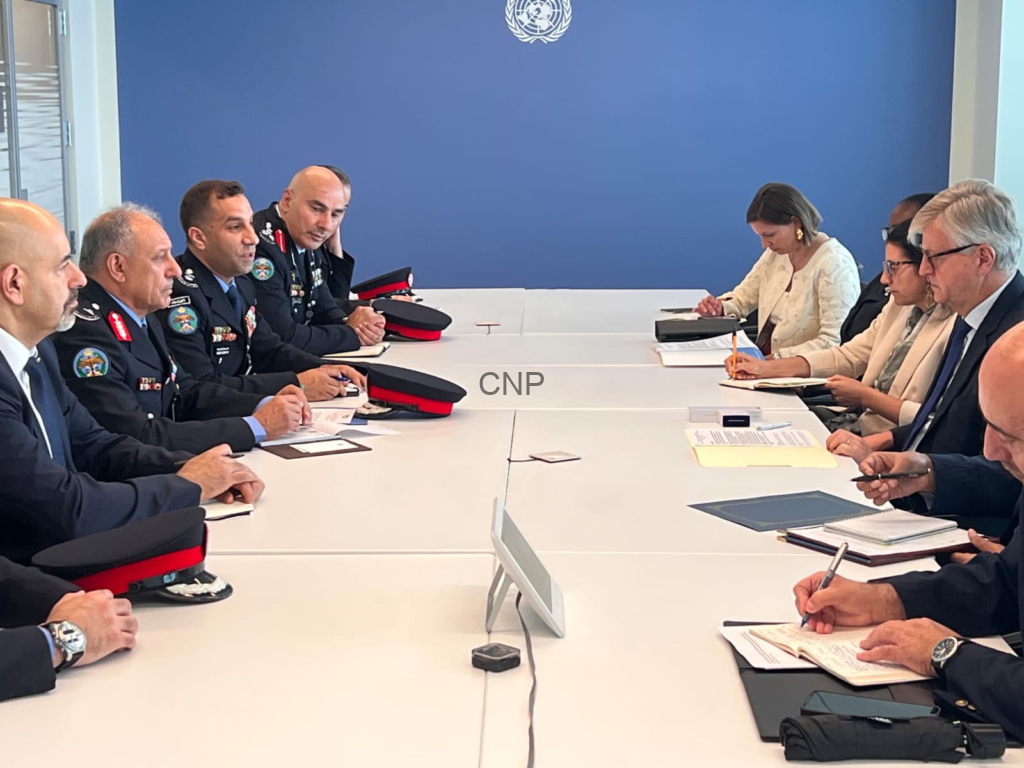 image - Center News Network Military officers and civilian diplomats sit around a long conference table in a United Nations meeting room, blue wall with UN emblem behind them, taking notes and discussing.