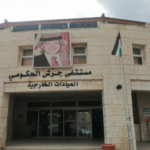 Exterior of Jerash Government Hospital with a portrait of the Jordanian king and Jordanian flags at the entrance; Arabic sign naming outpatient clinics.