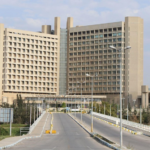 Front view of a large, multi-wing hospital building seen from a driveway leading to the main entrance with streetlights and landscaping.