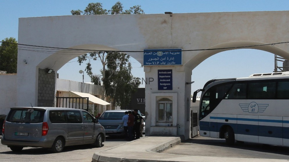 Border checkpoint entrance with a blue 'Arrivals and Departures' sign, a VIP desk, and a large bus on the right amid parked cars.