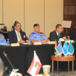 Panel of officials seated at a long table during a formal conference; uniformed officers in blue dress with medals flank suited delegates, microphones and nameplates visible.